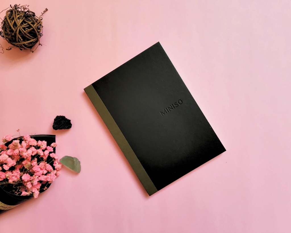 ピンクのデスクに黒のノートと花